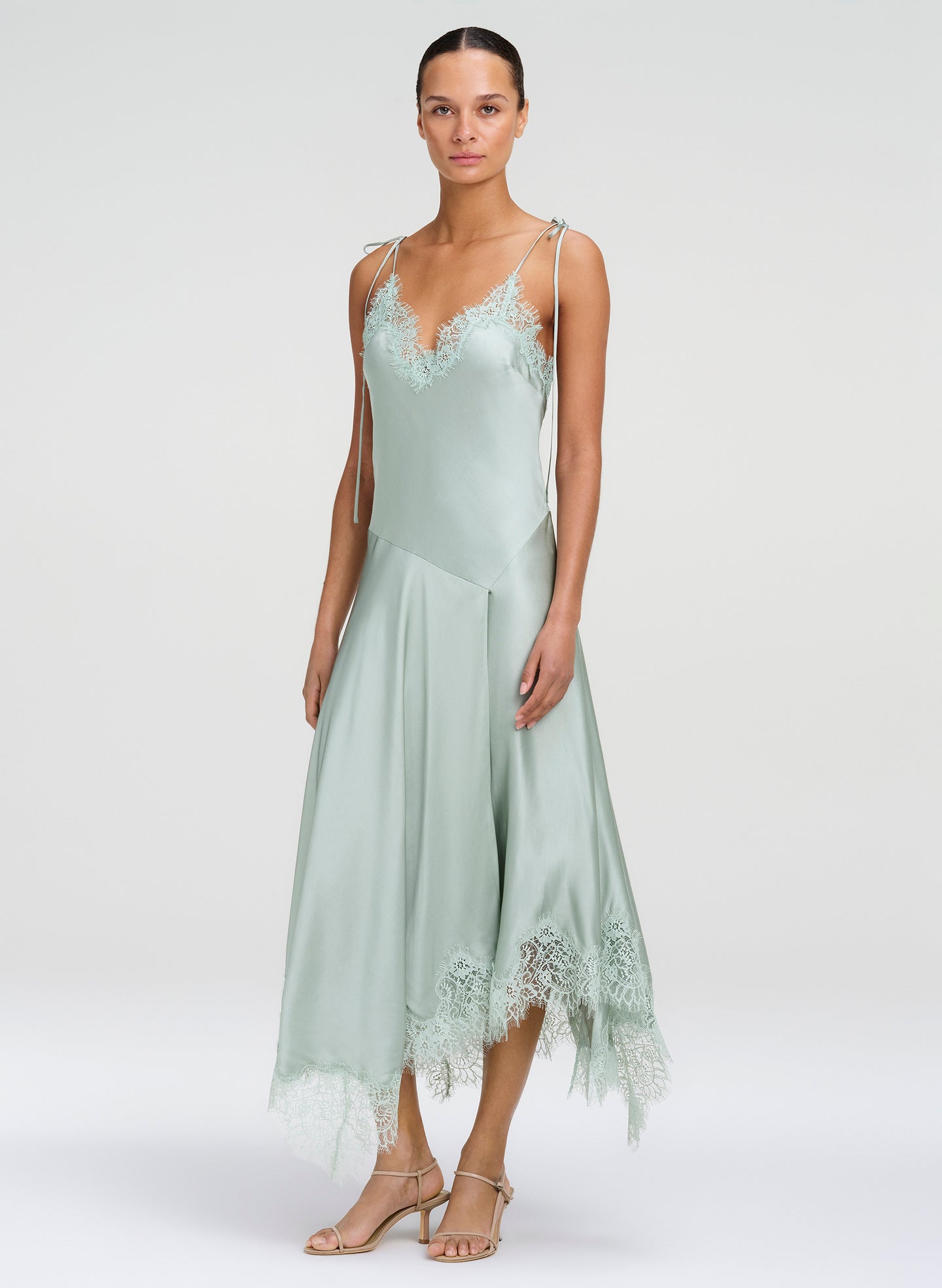 A model wearing a sage green satin slip dress with lace trim on a light gray studio background.