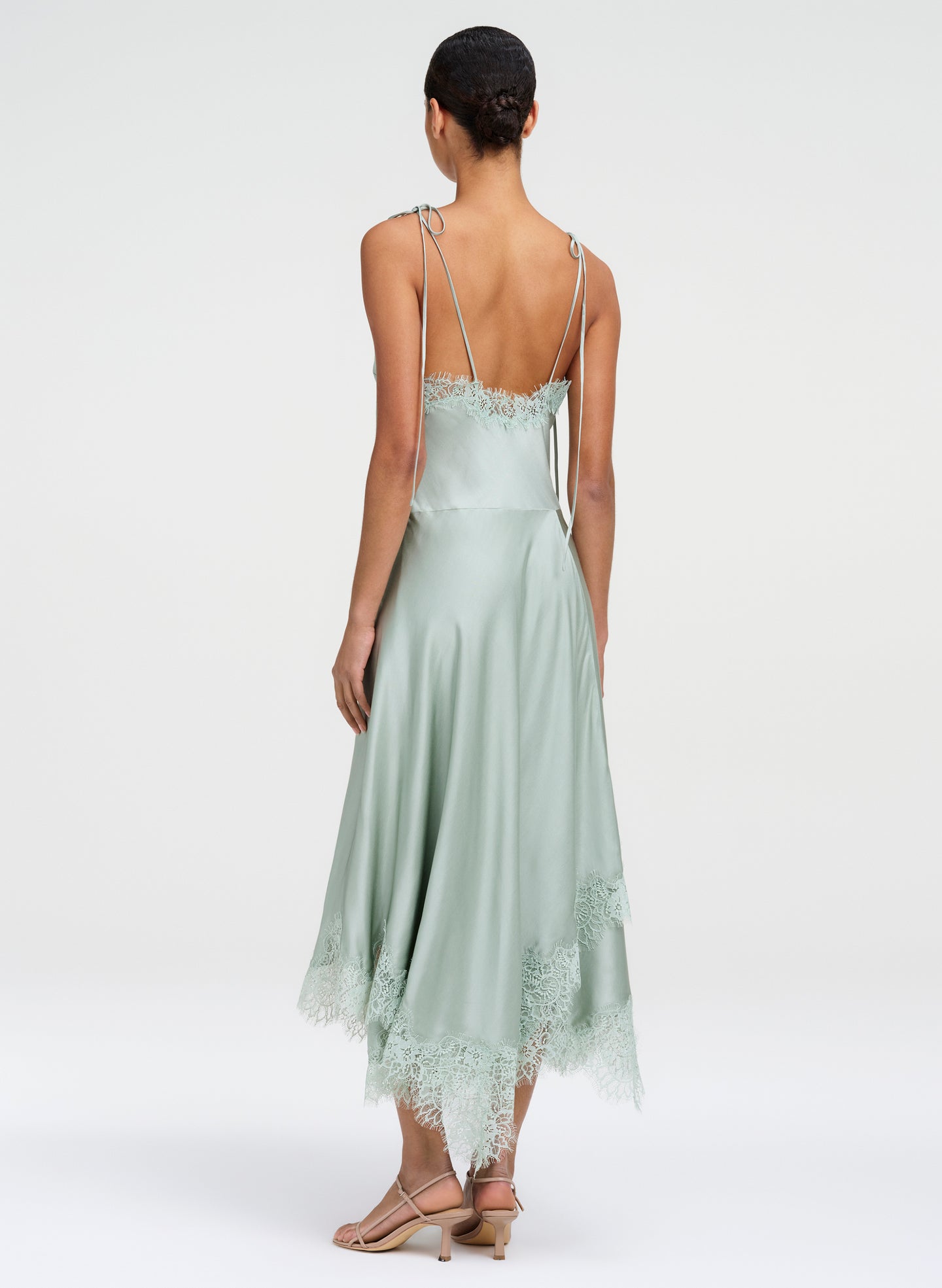 A woman shown from the back wearing a pale sage green satin midi dress with thin straps, lace trim at the neckline and hem, and a low back, photographed against a light gray studio background.
