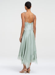 A woman shown from the back wearing a pale sage green satin midi dress with thin straps, lace trim at the neckline and hem, and a low back, photographed against a light gray studio background.