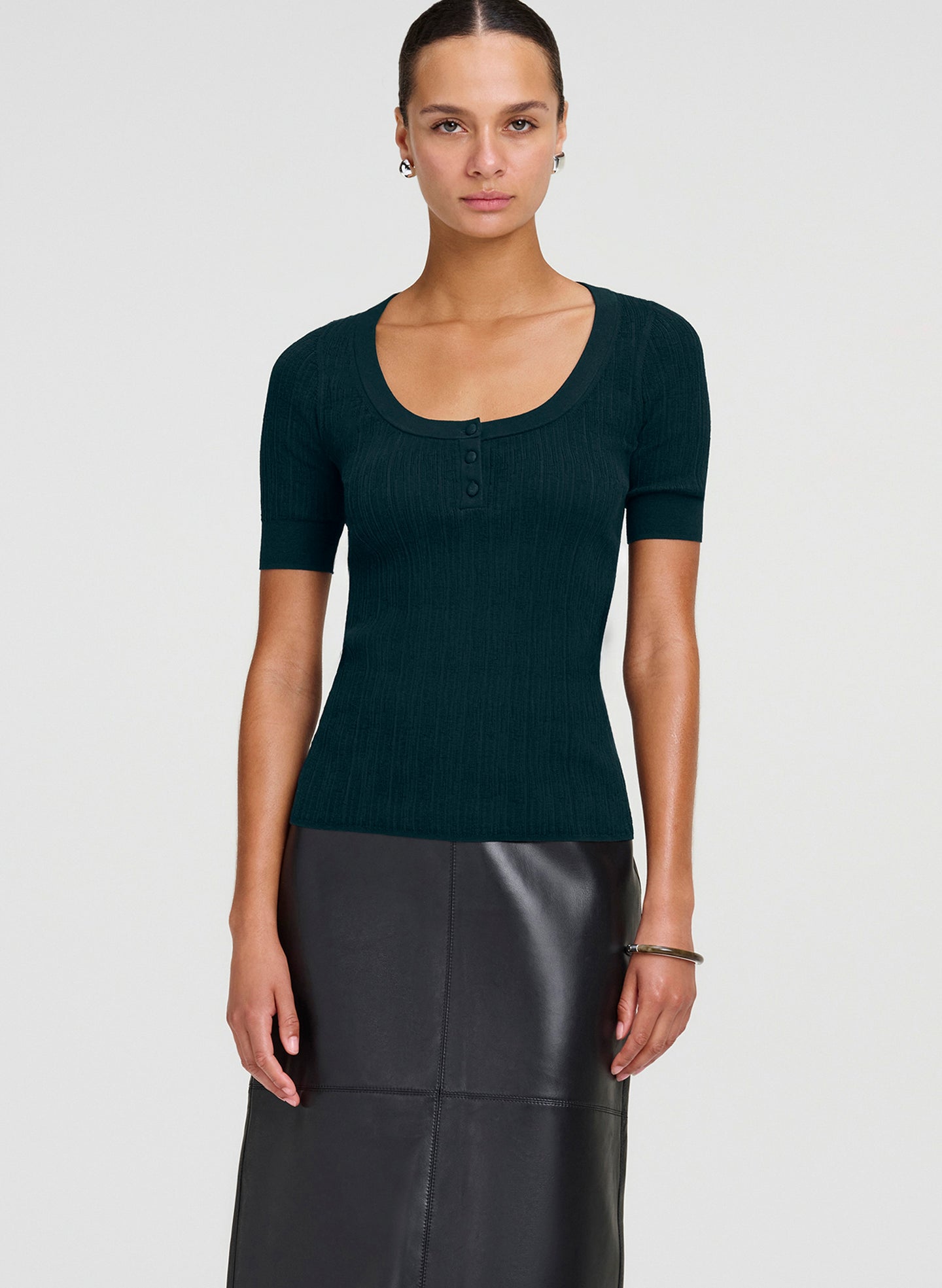A woman modeling a dark teal green short-sleeve ribbed knit top with a scoop neckline and three-button placket, paired with a black leather skirt, against a light gray studio background.