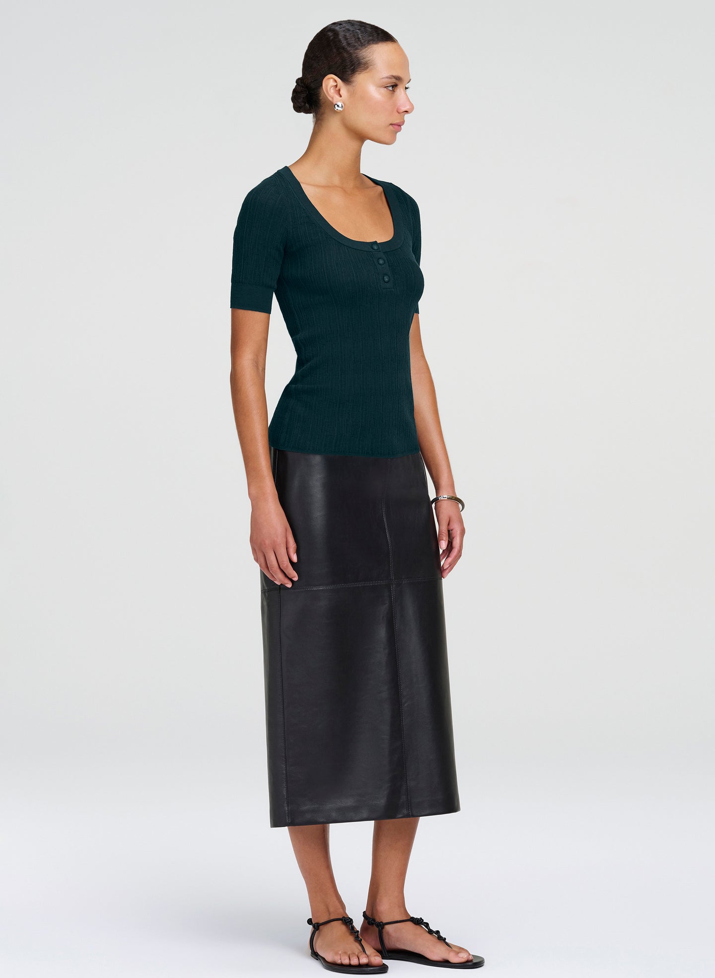 A woman modeling a dark green short-sleeve knit top and a black midi skirt, shown in profile against a light gray studio background.