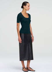 A woman modeling a dark green short-sleeve knit top and a black midi skirt, shown in profile against a light gray studio background.
