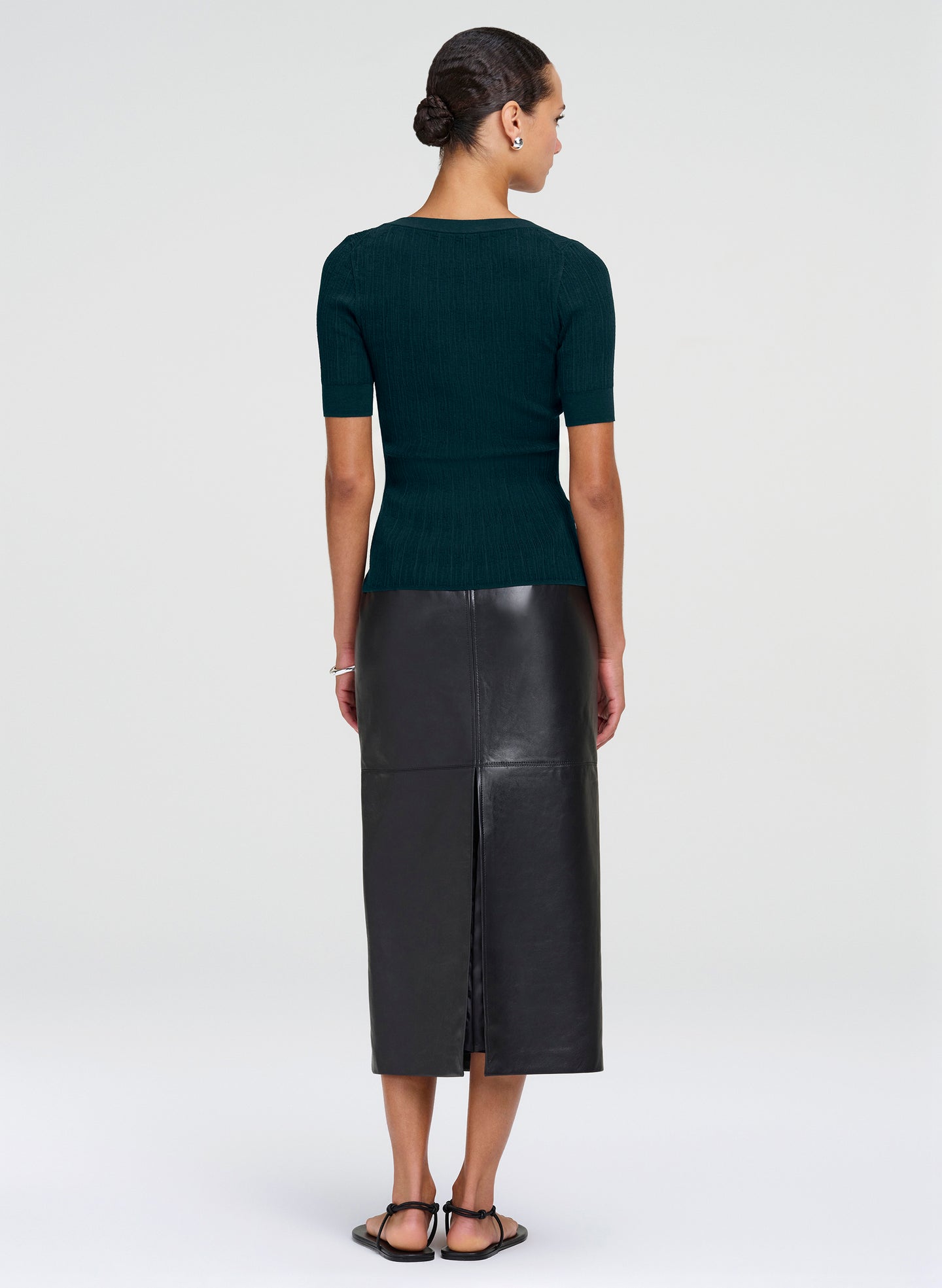 A woman shown from the back wearing a dark green short-sleeve ribbed knit top and a black leather midi skirt with a front slit, paired with black sandals, against a light gray studio background.