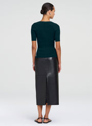 A woman shown from the back wearing a dark green short-sleeve ribbed knit top and a black leather midi skirt with a front slit, paired with black sandals, against a light gray studio background.