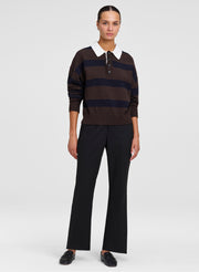 A model stands against a light gray studio background wearing a dark brown and navy horizontally striped knit polo sweater with a white collar, paired with black wide-leg trousers and black shoes.