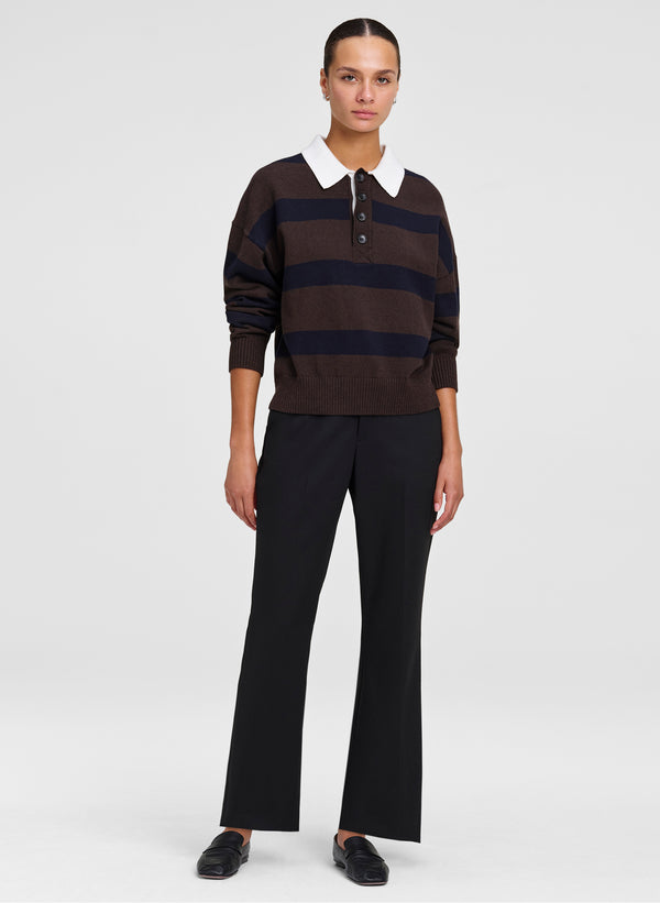 A model stands against a light gray studio background wearing a dark brown and navy horizontally striped knit polo sweater with a white collar, paired with black wide-leg trousers and black shoes.