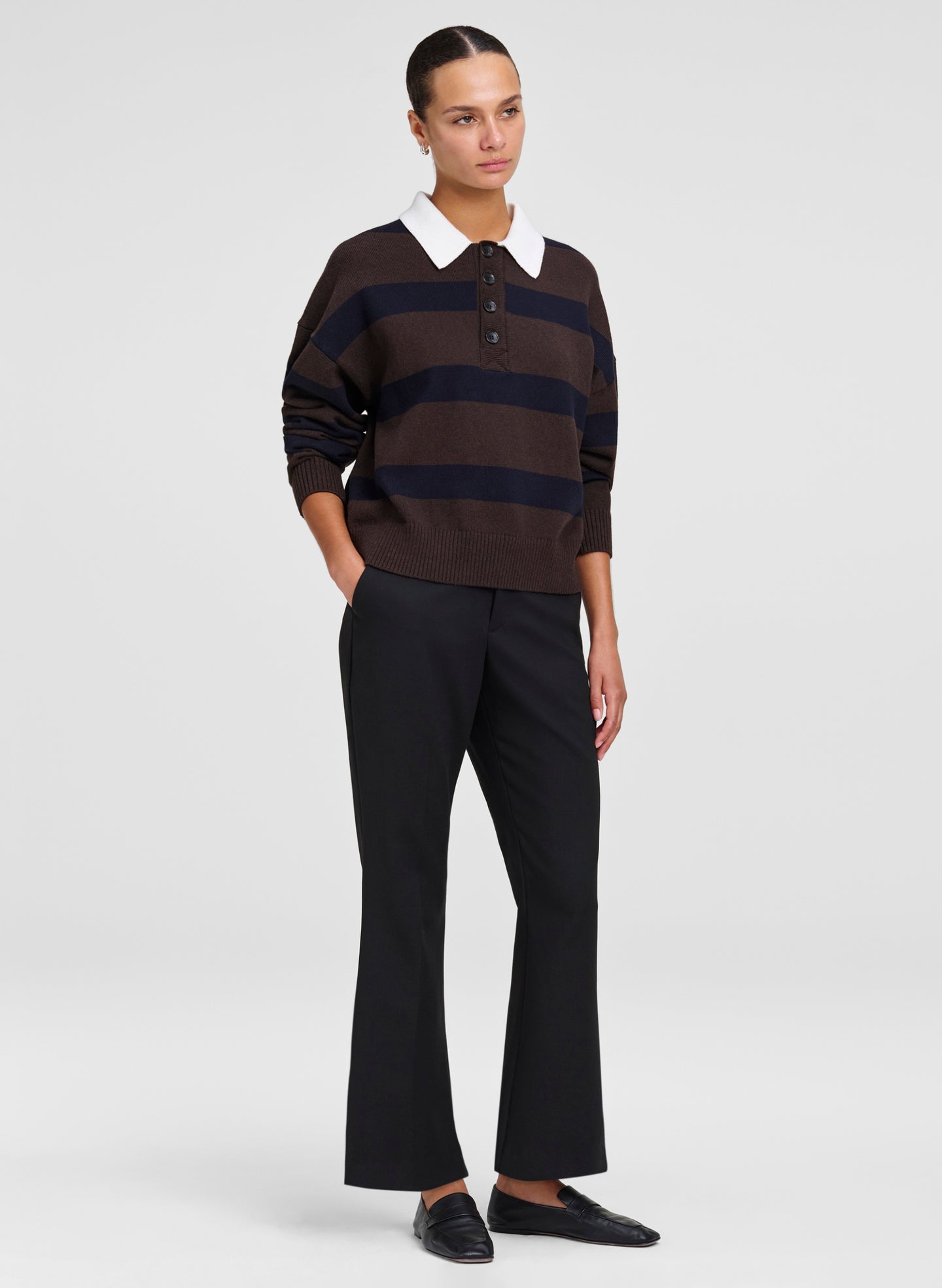 A model stands against a light gray studio background wearing a dark brown and navy horizontally striped knit polo sweater with a white collar, black flared trousers, and black loafers.