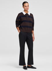 A model stands against a light gray studio background wearing a dark brown and navy horizontally striped knit polo sweater with a white collar, black flared trousers, and black loafers.