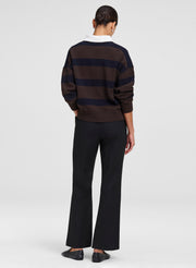A model shown from the back wearing a dark brown and navy horizontally striped sweater over a white collared shirt, paired with black wide-leg trousers and dark loafers, against a light gray studio background.
