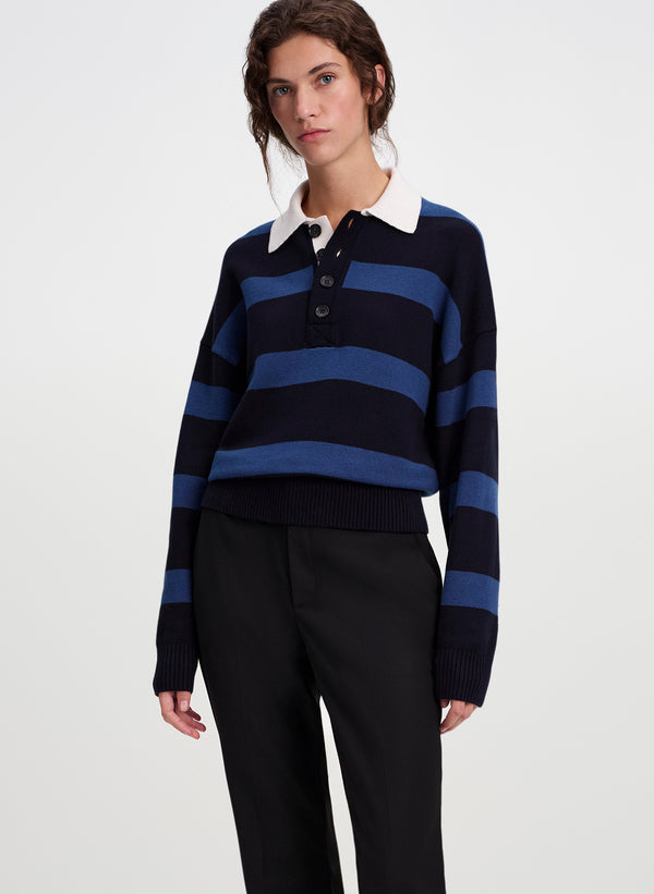 A model wearing a navy and blue horizontally striped knit polo sweater with a white collar, paired with black trousers, against a plain light gray studio background.