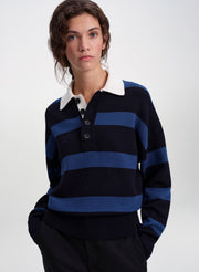 A woman wearing a navy and blue horizontally striped knit polo sweater with a white collar, paired with dark trousers, against a light gray studio background.