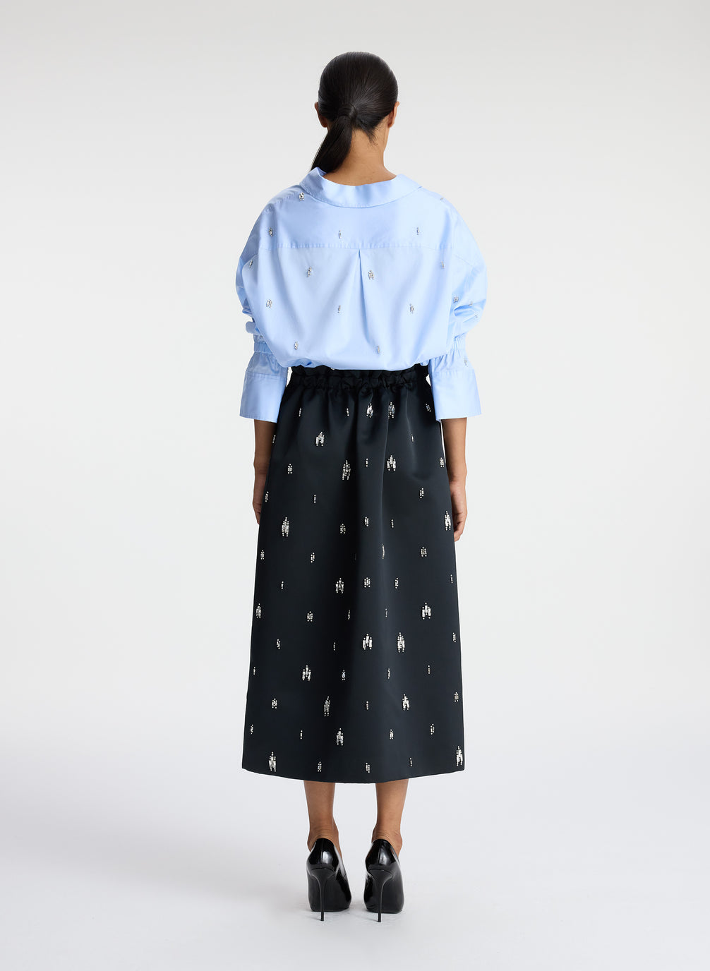 back view of woman wearing blue embellished button down top and black embellished midi skirt