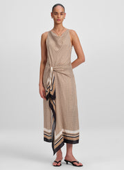 A model wearing a sleeveless, wrap-style midi dress with a beige and black geometric pattern and a striped border, paired with black slide sandals, against a light gray studio background.