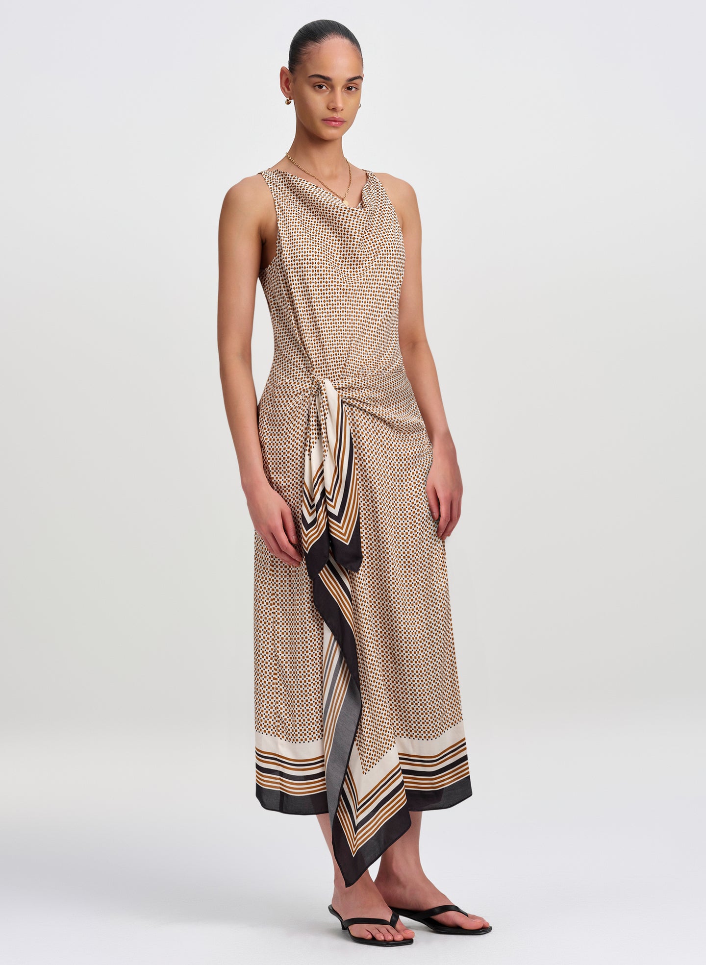 A model wears a sleeveless, cowl-neck midi dress with a beige, black, and white geometric pattern and a draped side panel. The dress features a tie waist and a striped border at the hem, styled with black flip-flops against a light gray studio background.