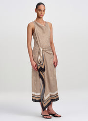 A model wears a sleeveless, cowl-neck midi dress with a beige, black, and white geometric pattern and a draped side panel. The dress features a tie waist and a striped border at the hem, styled with black flip-flops against a light gray studio background.