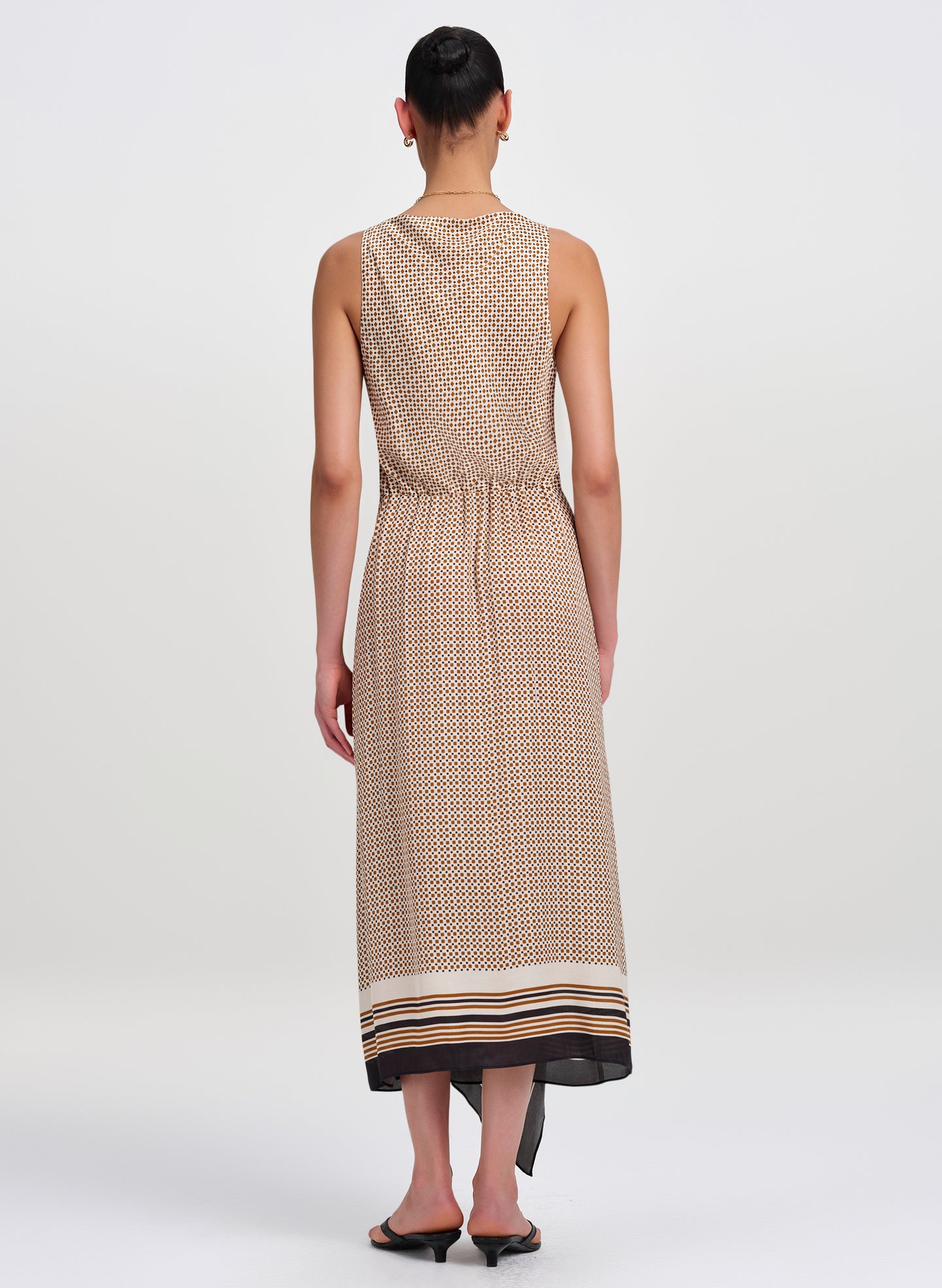 A woman viewed from the back wearing a sleeveless midi dress with a beige and brown geometric pattern and a black hem, paired with black flat sandals, against a light gray studio background.