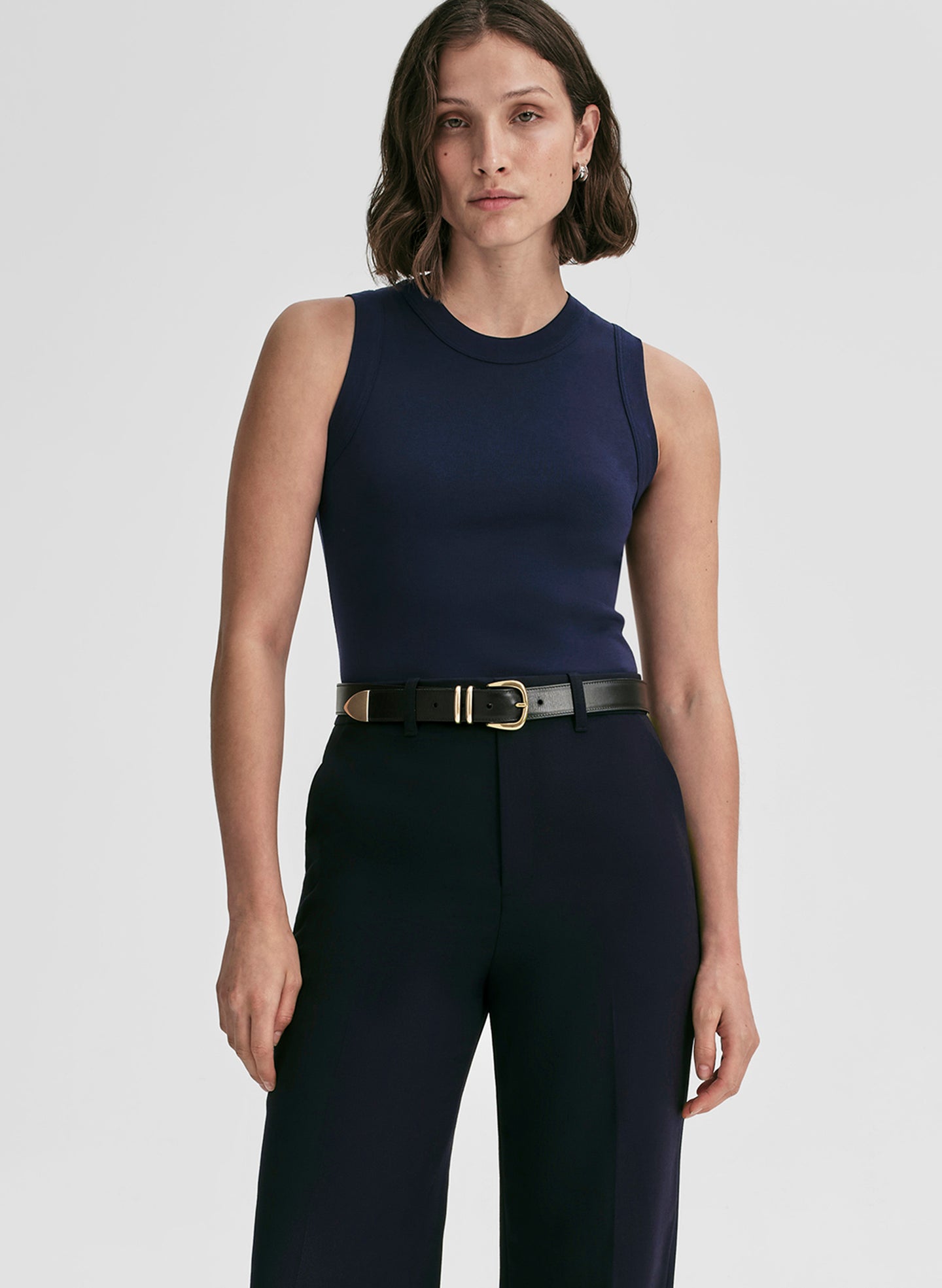 Woman wearing a navy sleeveless top and pants with a gold belt on a white background