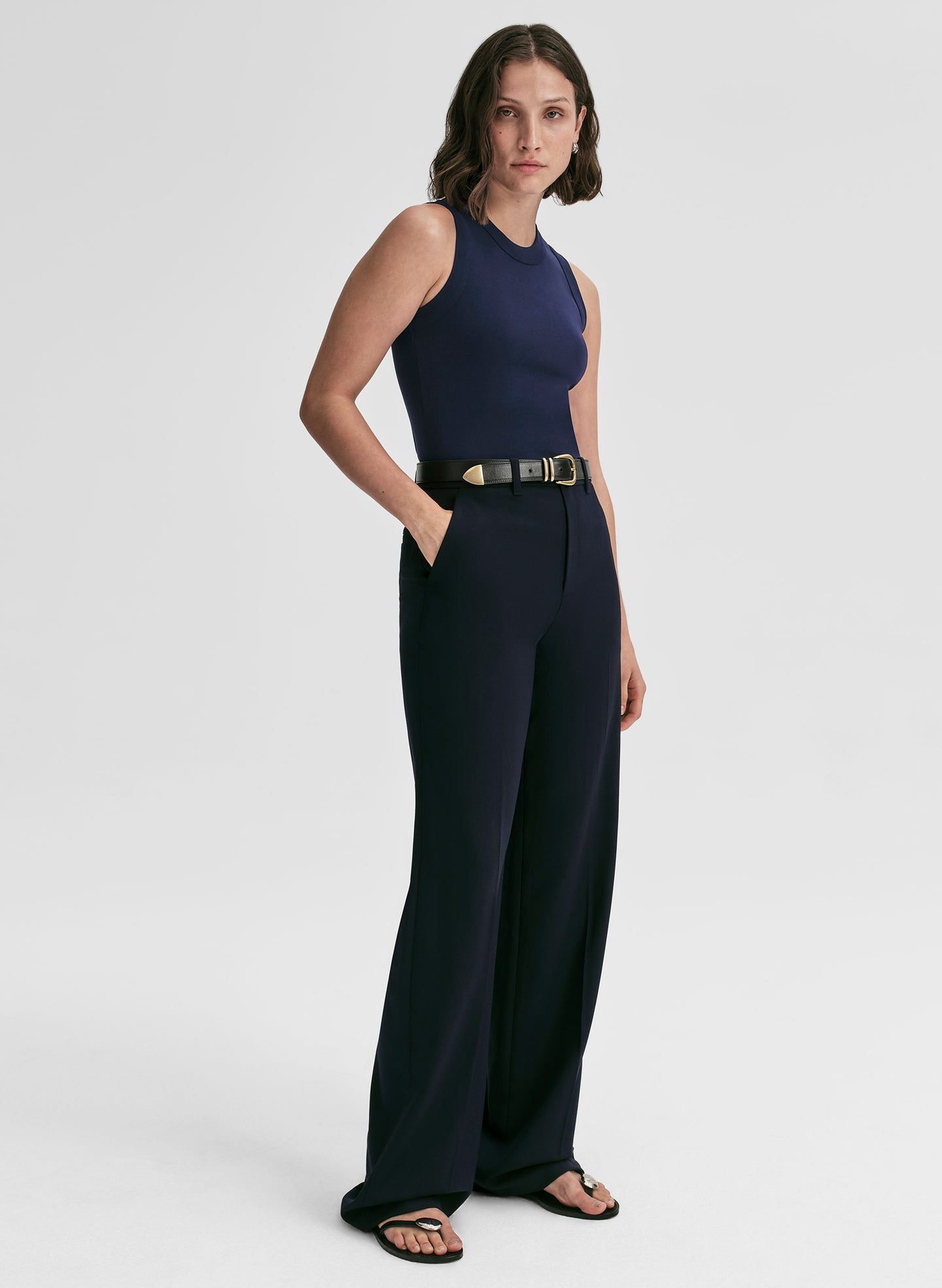 Woman wearing a navy sleeveless top and pants with a gold belt on a white background