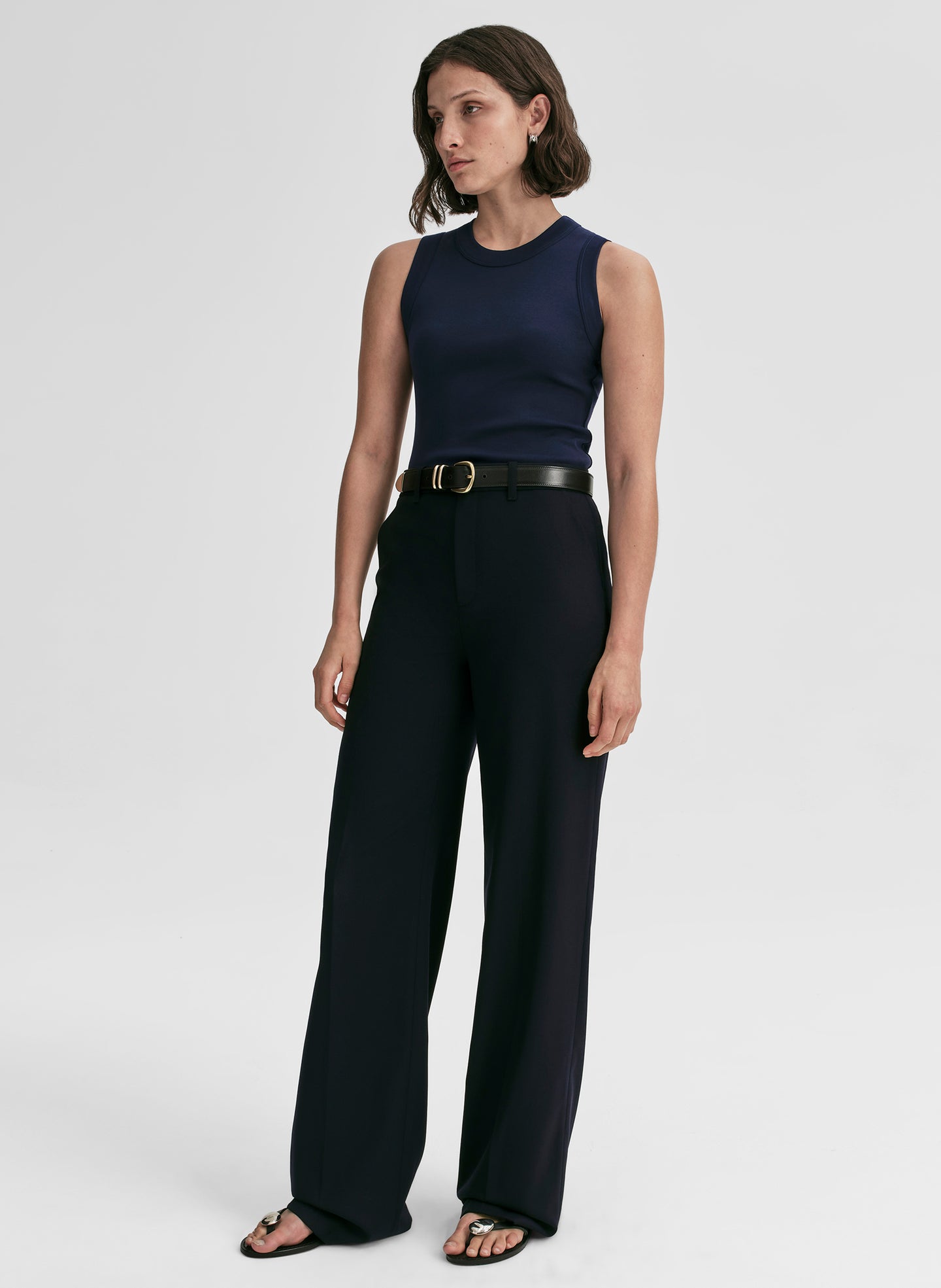 Woman wearing a navy sleeveless top and pants with a gold belt on a white background