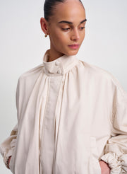 A close-up of a model wearing a light beige high-neck jacket with gathered ruffles and button details on the collar, set against a plain light gray background.