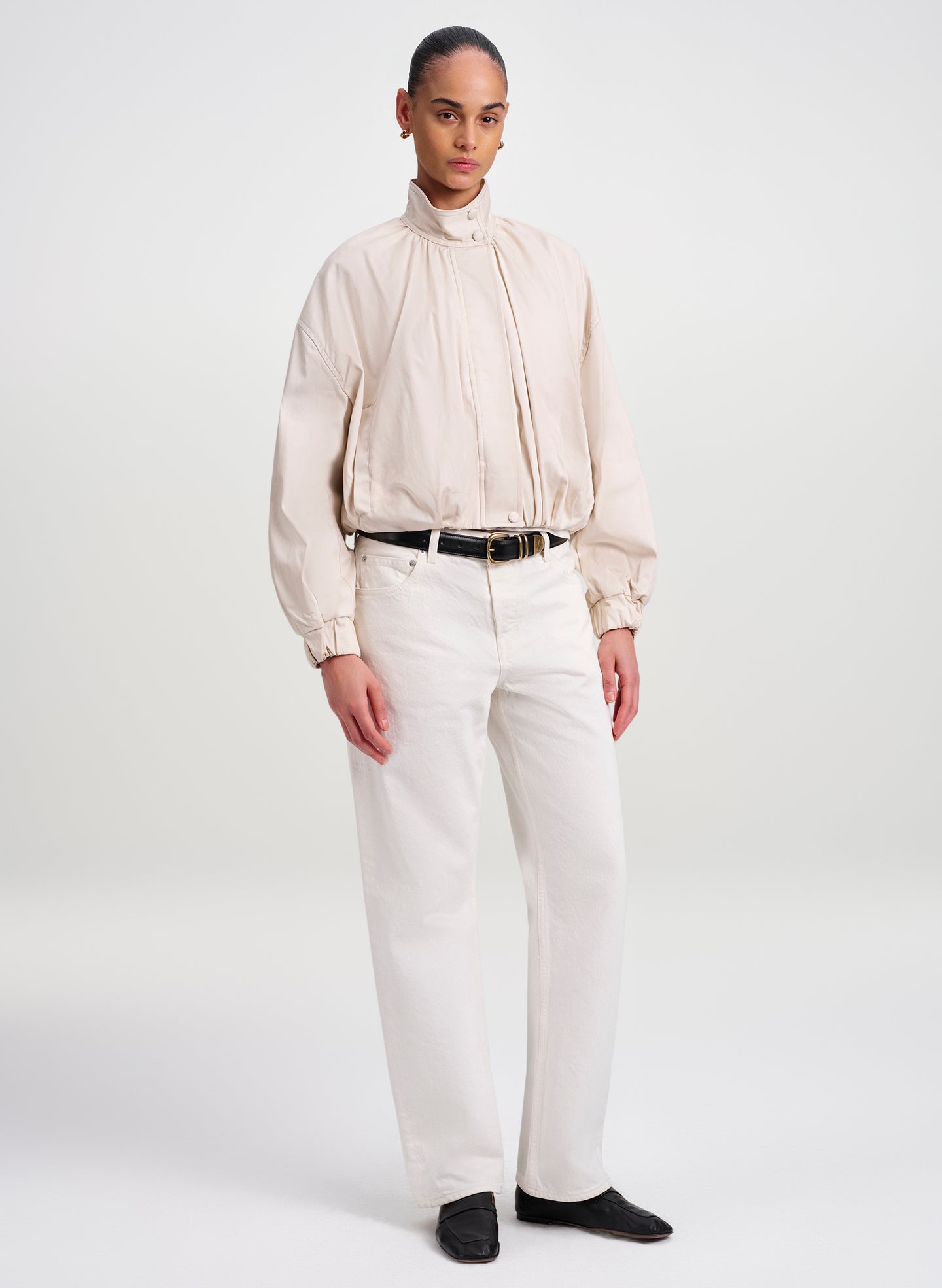 A model stands against a light gray studio background wearing a cream-colored high-neck long-sleeve jacket with gathered detailing, paired with off-white trousers, a black belt, and black shoes.