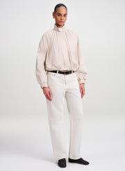 A model stands against a light gray studio background wearing a cream-colored high-neck long-sleeve jacket with gathered detailing, paired with off-white trousers, a black belt, and black shoes.