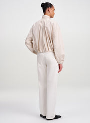 A model shown from the back wearing a light beige high-neck long-sleeve jacket  and off-white wide-leg trousers, paired with black shoes, against a plain light gray studio background.