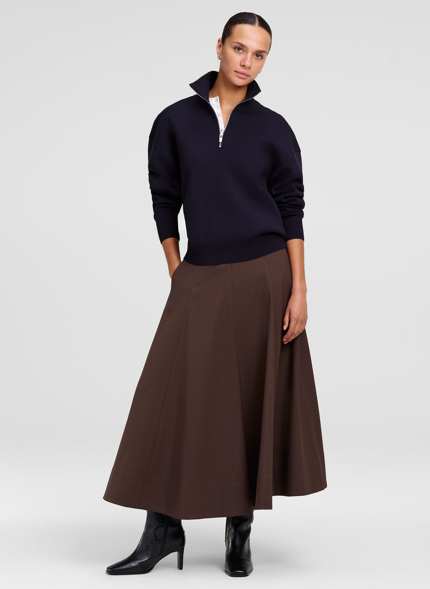 A model stands against a light gray studio background wearing a dark navy quarter-zip sweater over a white shirt, paired with a brown A-line midi skirt and black boots.