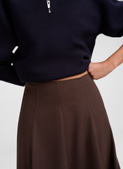 Close-up of a person wearing a dark navy quarter-zip top and a brown skirt, cropped from the chest down against a light gray studio background.