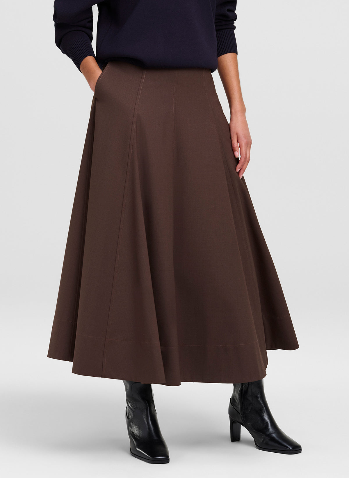 A woman models a dark brown A-line midi skirt with side pockets, paired with a navy long-sleeve top and black boots, against a light gray studio background.