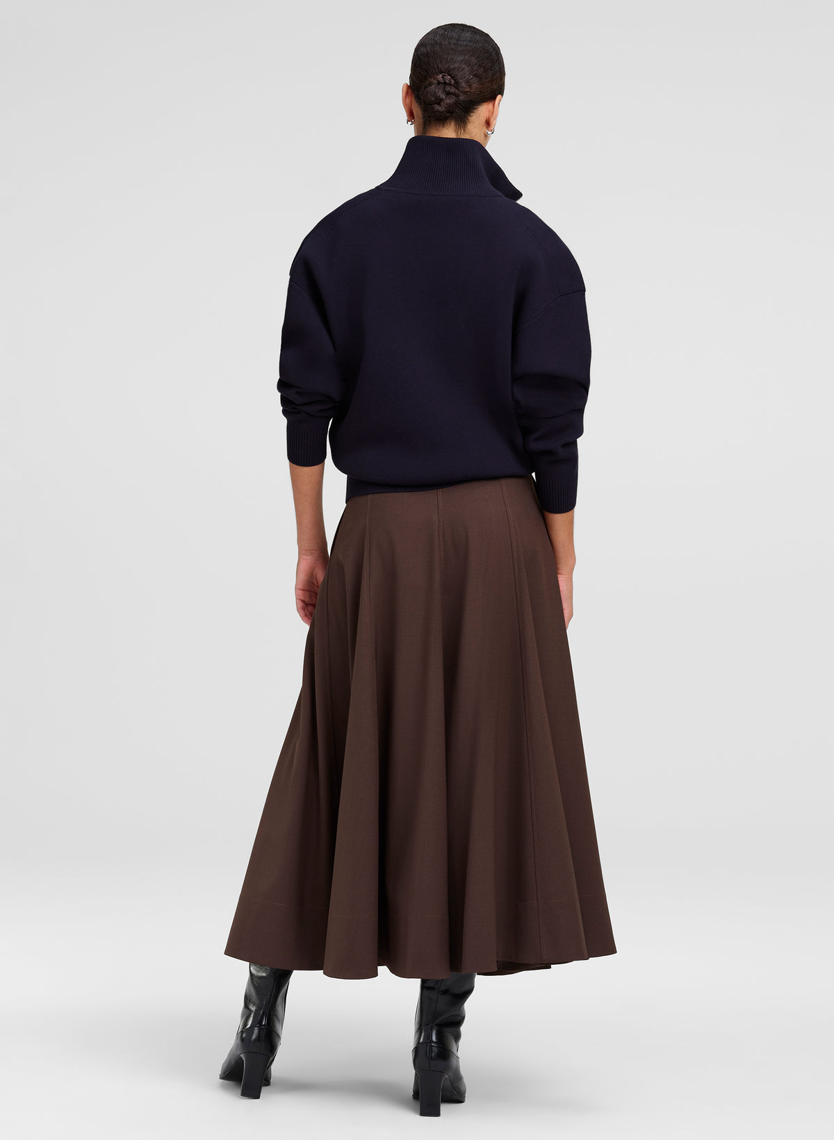 A model shown from the back wearing a dark navy mock-neck knit sweater and a brown A-line midi skirt, paired with black  boots, against a light gray studio background.