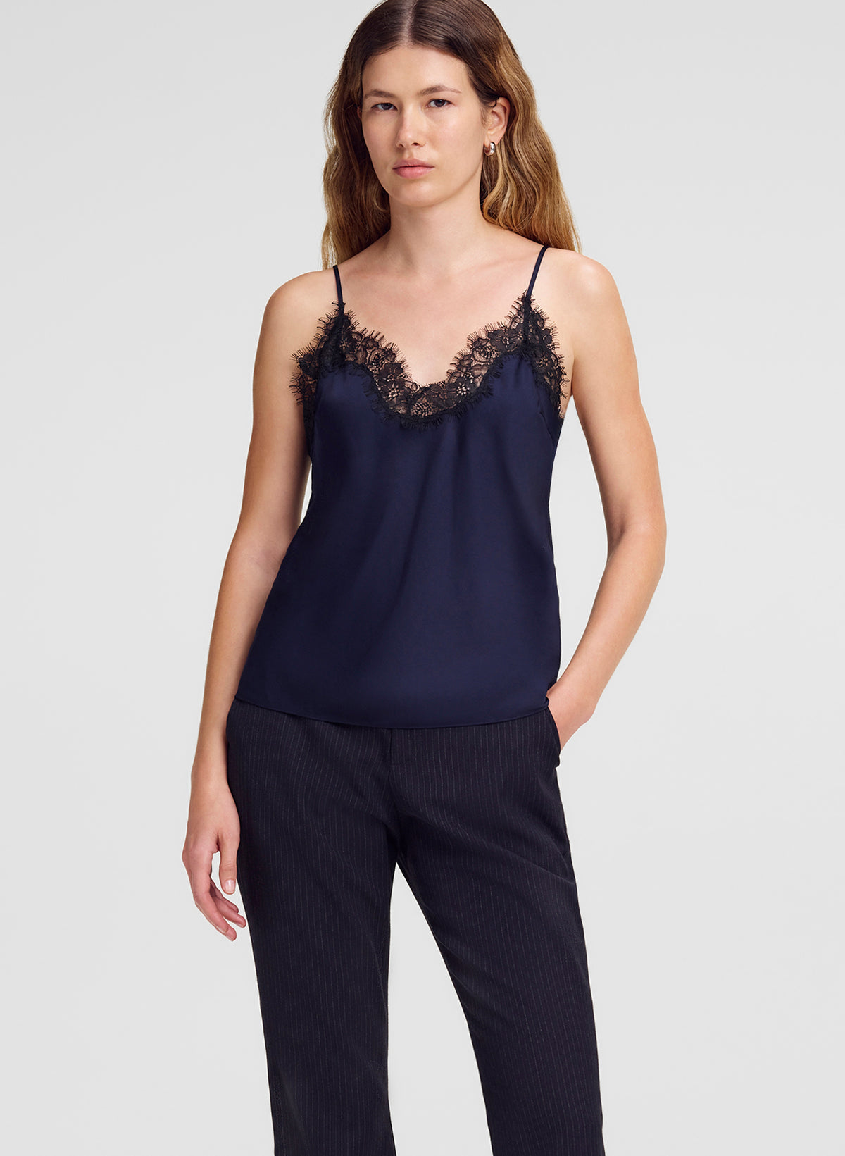 A woman modeling a navy blue camisole top with black lace trim along the neckline, paired with dark pinstriped trousers, against a light gray studio background.