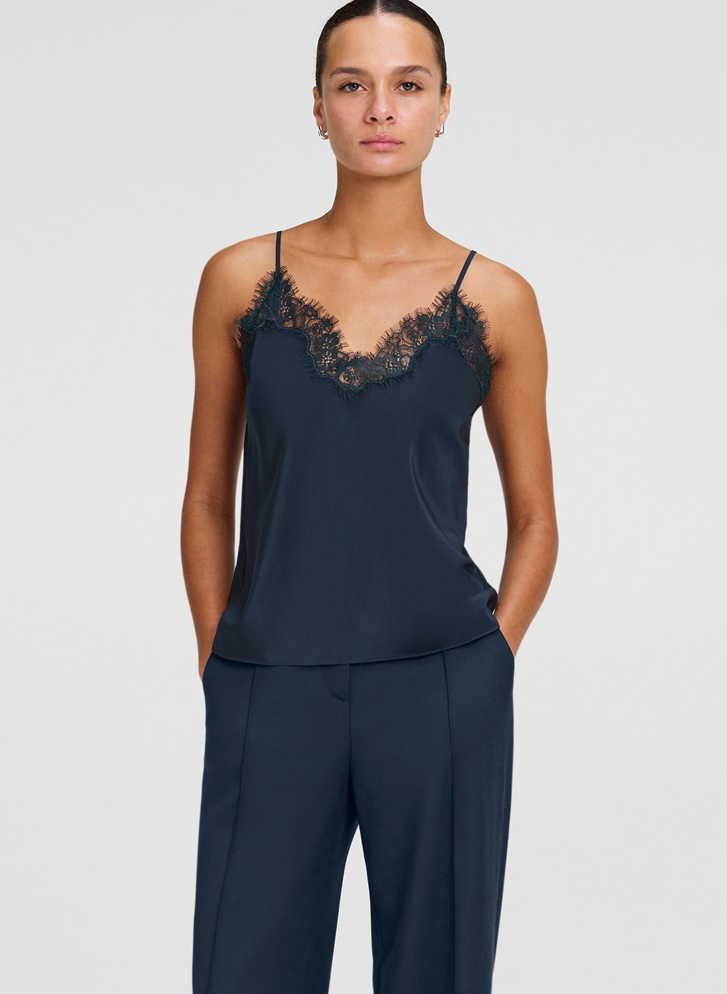 A model wearing a dark green camisole top with black lace trim at the neckline, paired with matching navy trousers, against a light gray studio background.