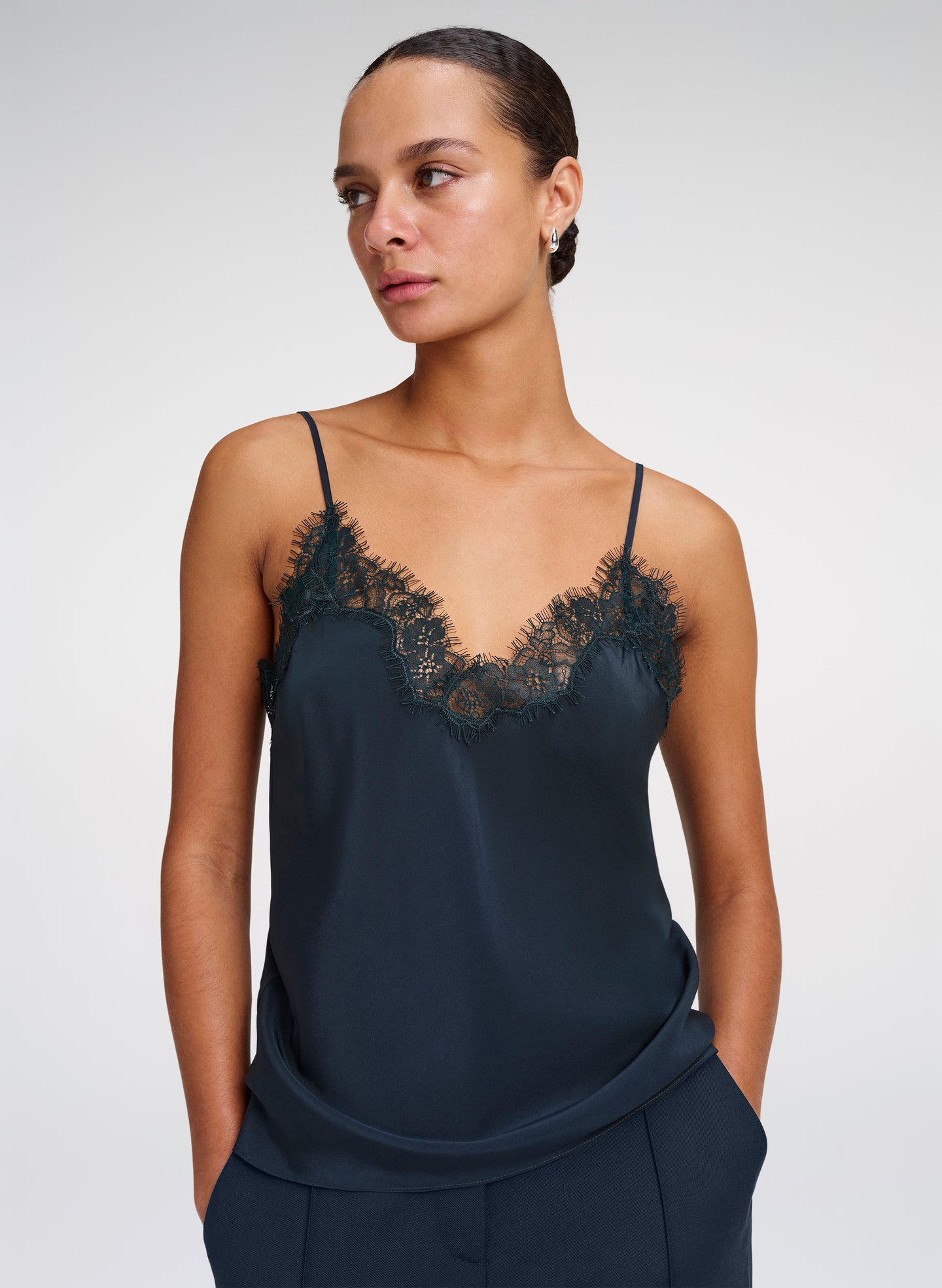A woman modeling a dark green satin camisole top with black lace trim along the V-neckline, paired with matching green trousers, against a light gray studio background.