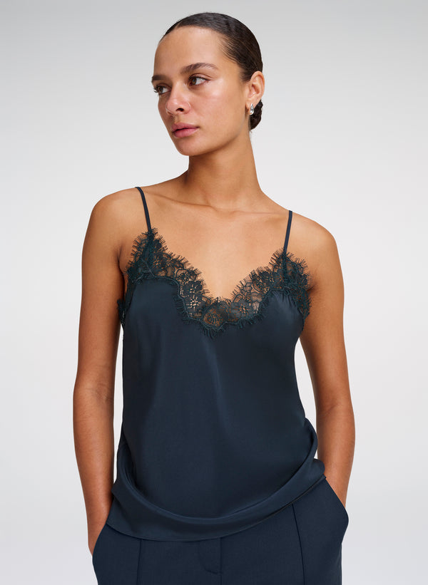 A woman modeling a dark green satin camisole top with black lace trim along the V-neckline, paired with matching green trousers, against a light gray studio background.