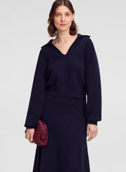 A woman modeling a dark navy blue long-sleeve sweater with a V-neckline and draped shoulder detail, holding a small textured burgundy clutch against a light gray studio background.
