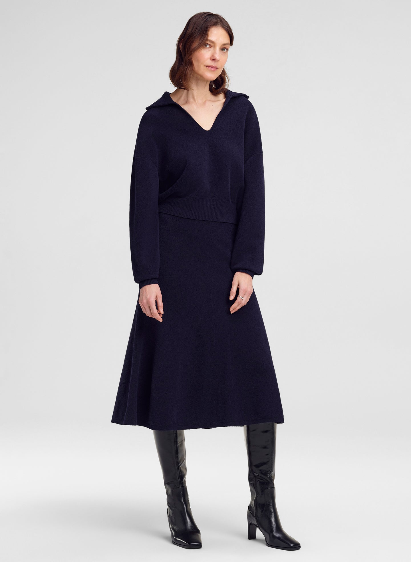 A woman modeling a dark navy collared long-sleeve sweater with a V-neck and flared skirt, paired with black knee-high boots, against a light gray studio background.