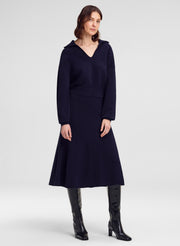 A woman modeling a dark navy collared long-sleeve sweater with a V-neck and flared skirt, paired with black knee-high boots, against a light gray studio background.