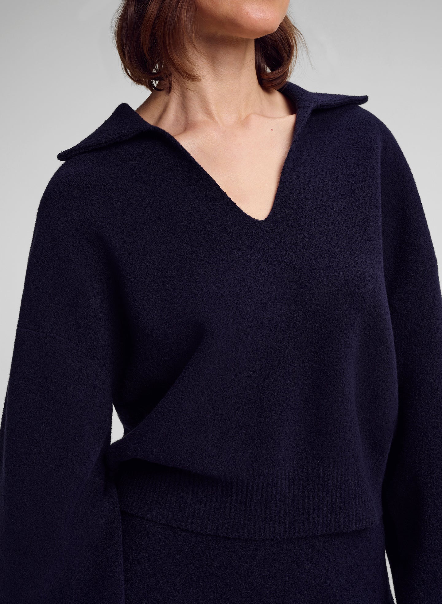 A close-up of a model wearing a dark navy blue knit sweater with a wide collar and V-neckline, shown against a neutral gray background.