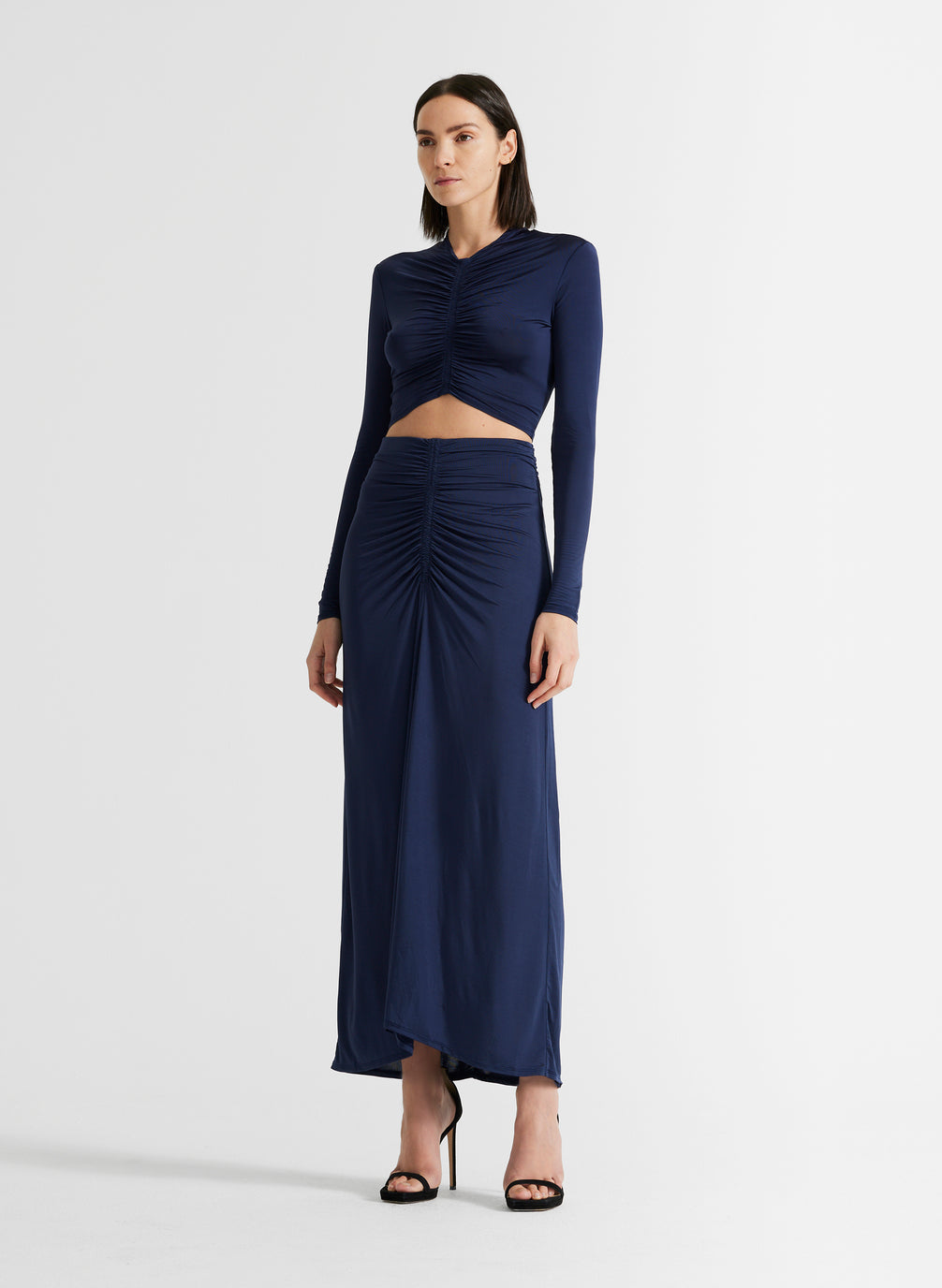 side view of woman wearing navy blue ruched long sleeve crop top and matching navy blue skirt