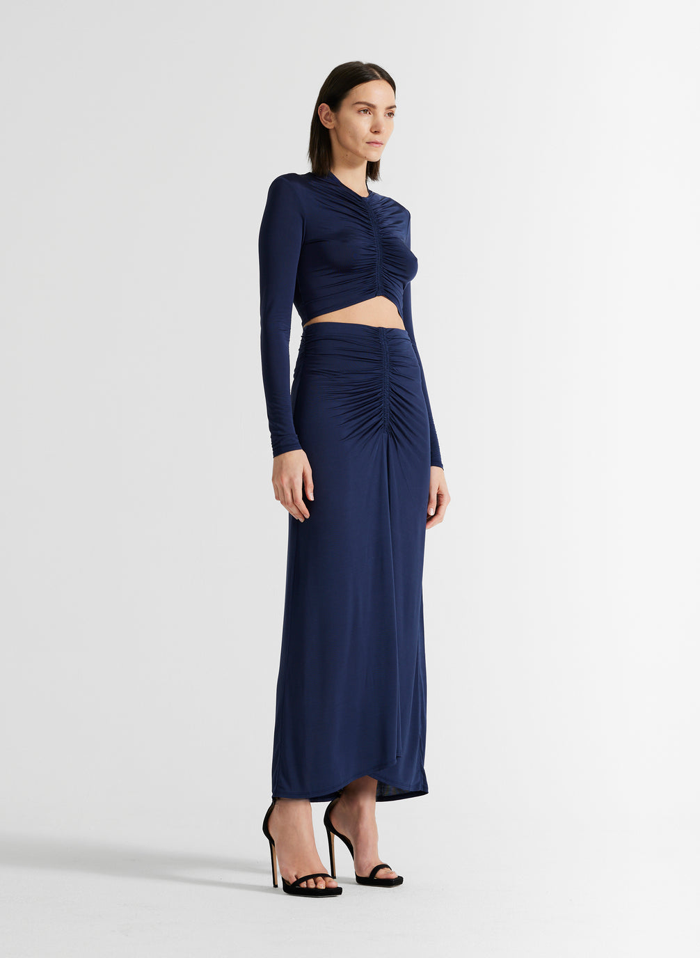 side view of woman wearing navy blue ruched long sleeve crop top and matching navy blue skirt