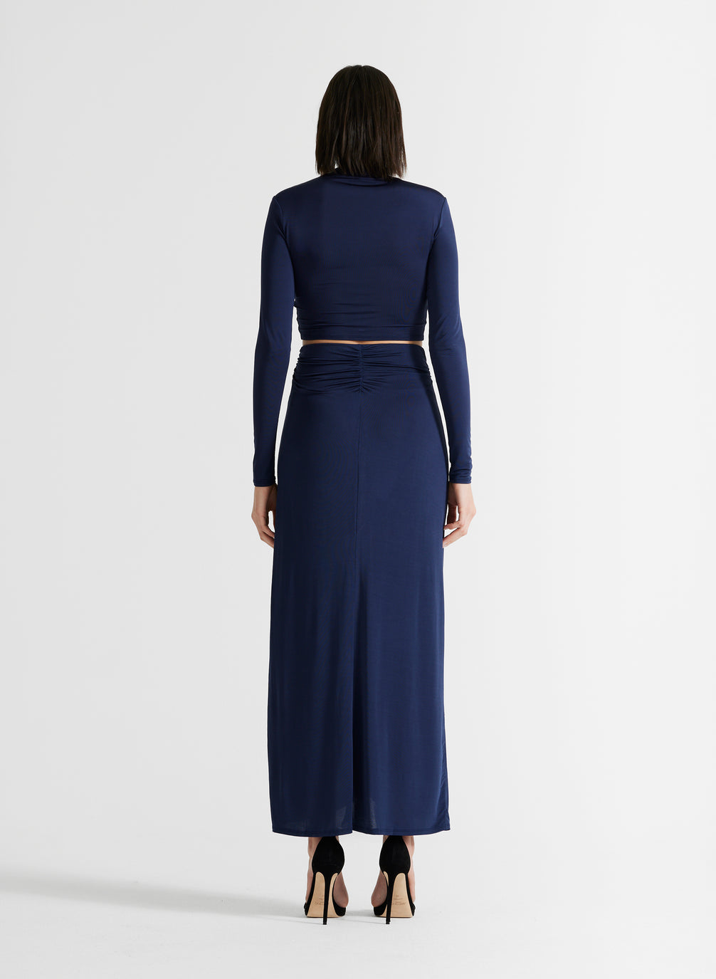 back view of woman wearing navy blue ruched long sleeve crop top and matching navy blue skirt