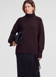 A woman modeling an oversized dark brown ribbed turtleneck sweater, paired with black trousers and holding a black clutch bag against a light gray studio background.