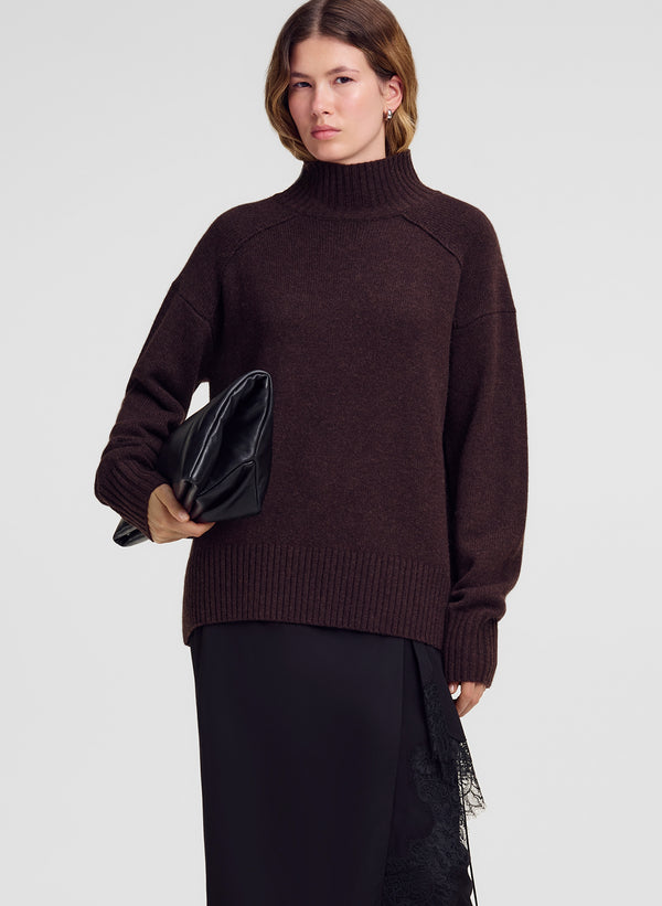 A woman modeling an oversized dark brown ribbed turtleneck sweater, paired with black trousers and holding a black clutch bag against a light gray studio background.