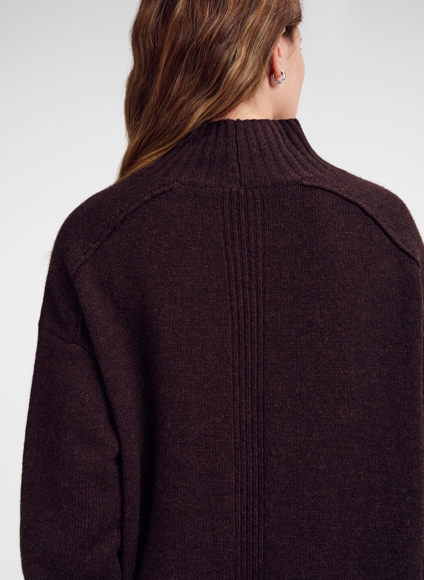 A close-up back view of a woman wearing a dark brown ribbed knit sweater against a light gray studio background.
