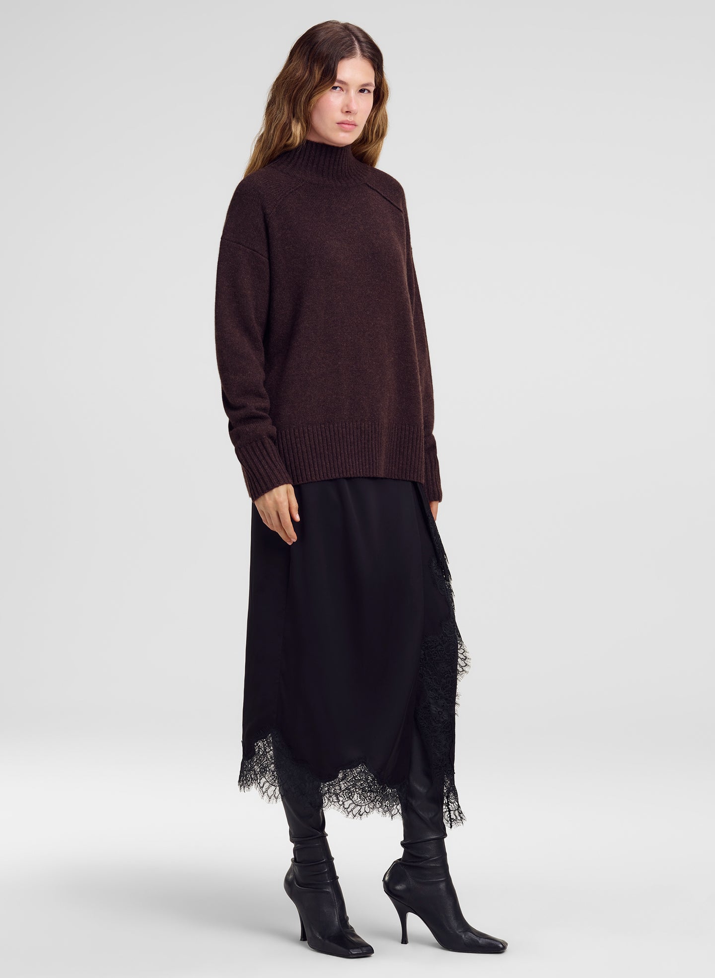 A model wearing a dark brown mock-neck knit sweater, a long black skirt with lace trim, black leggings, and black heeled boots, photographed against a light gray studio background.