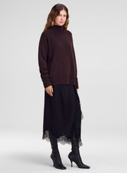 A model wearing a dark brown mock-neck knit sweater, a long black skirt with lace trim, black leggings, and black heeled boots, photographed against a light gray studio background.