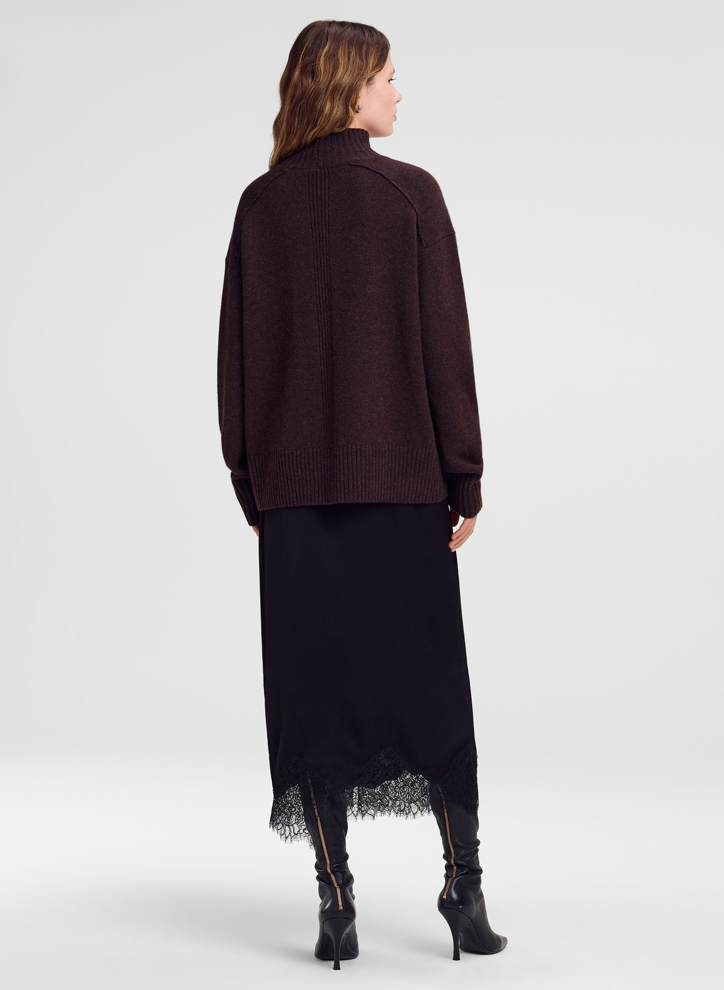 A woman shown from the back wearing a dark brown ribbed knit mock-neck sweater, a long black skirt with lace trim, and black heeled boots against a light gray studio background.