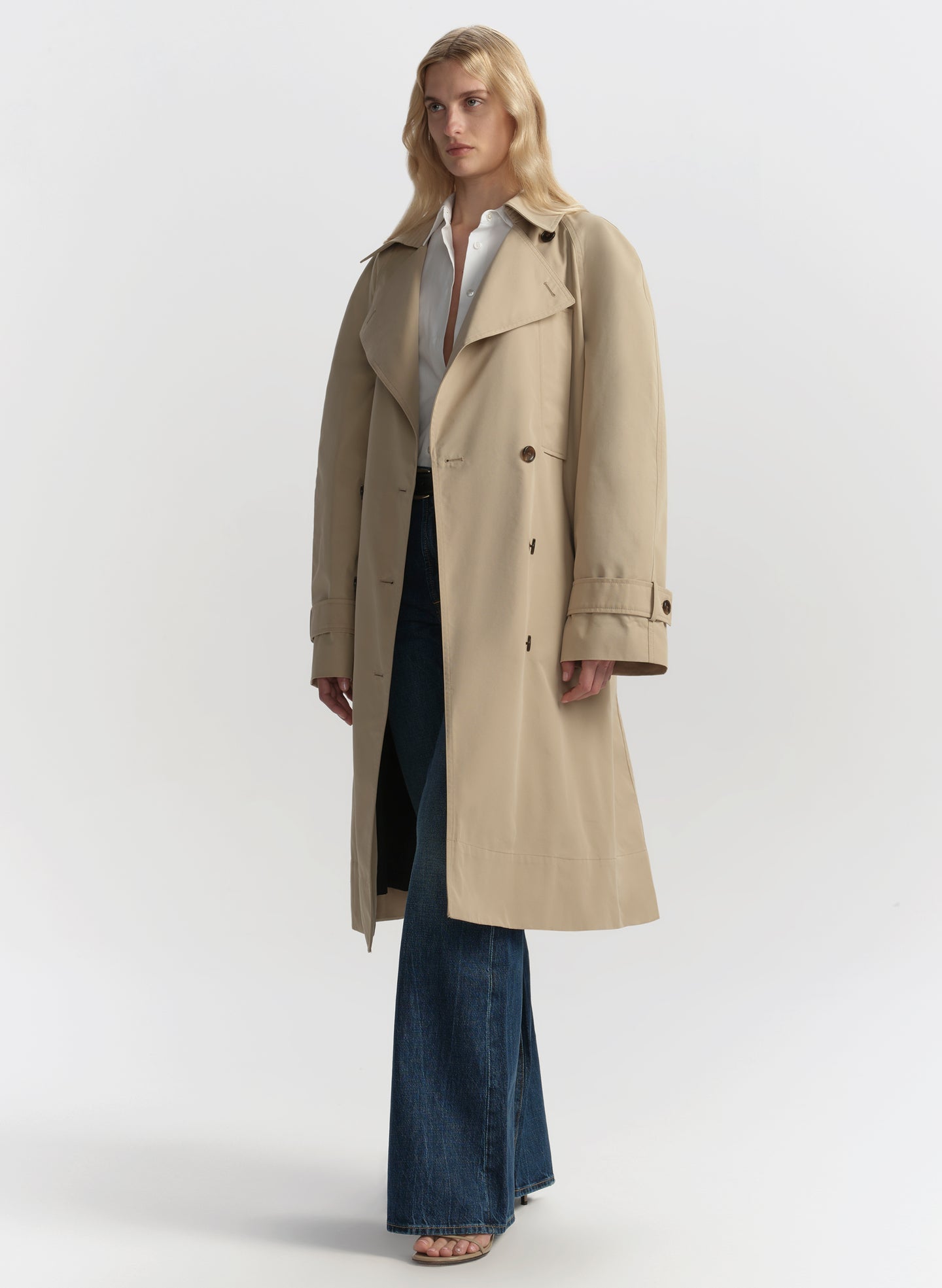 Woman wearing a beige trench coat, white shirt, and blue jeans on a plain background