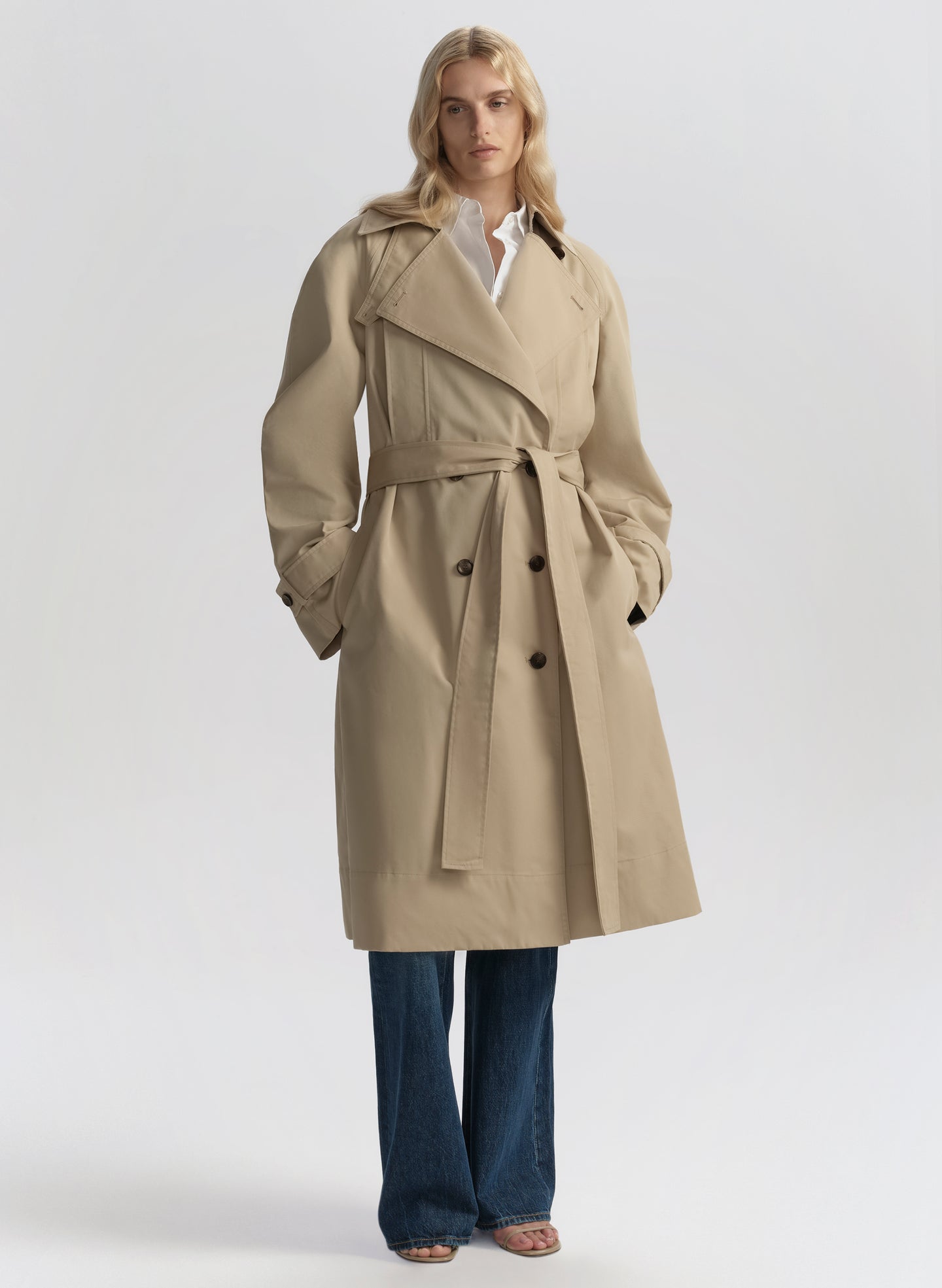 Woman wearing a beige trench coat, white shirt, and blue jeans on a plain background